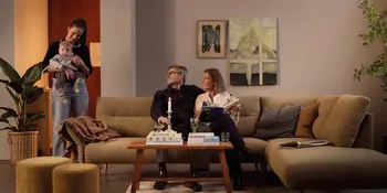 Frau mit Baby neben Sofa, zwei Personen sitzen auf Ecksofa mit Couchtisch und Kerze